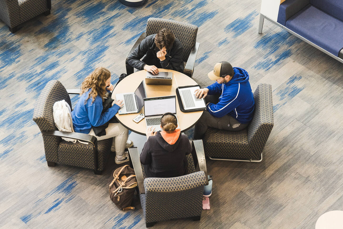 Four individuals are seated around a round table in a modern lounge, each focused on their own laptop.